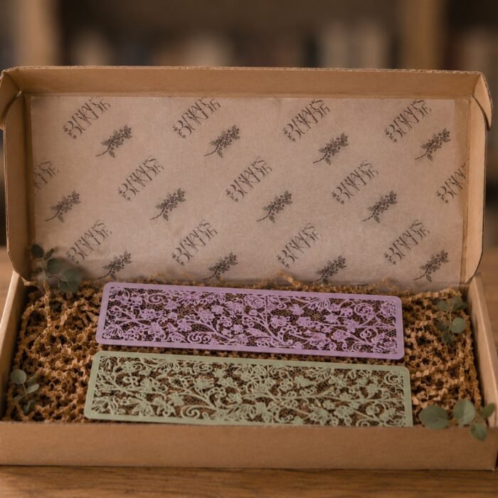 Dos marcapáginas calados imitando encaje floral, uno lila y otro verde salvia, sobre viruta de papel dentro de una caja